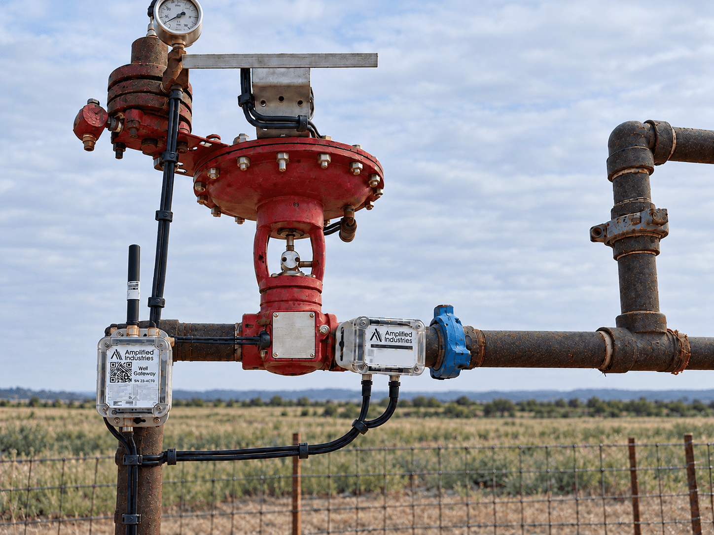 Two Amplified devices on a plunger lift wellhead in soft daylight, with a centered antenna on the left device
